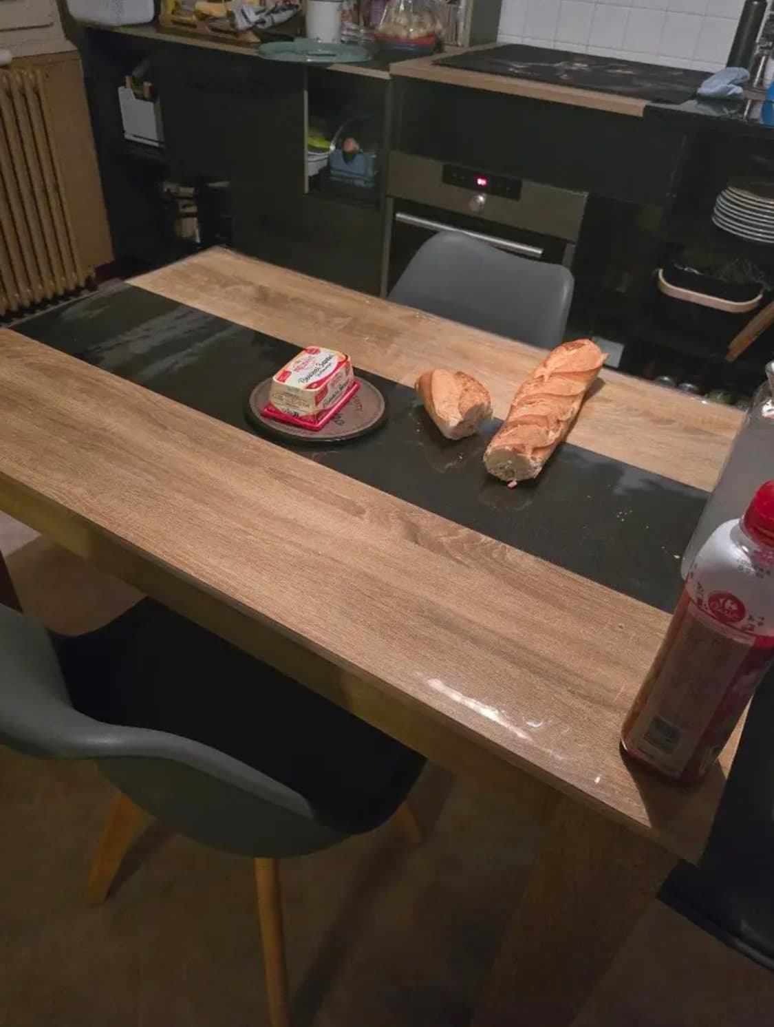 Keukentafel met een donkere strook in het midden, daarop stokbrood en boter; het blad onder zacht glas