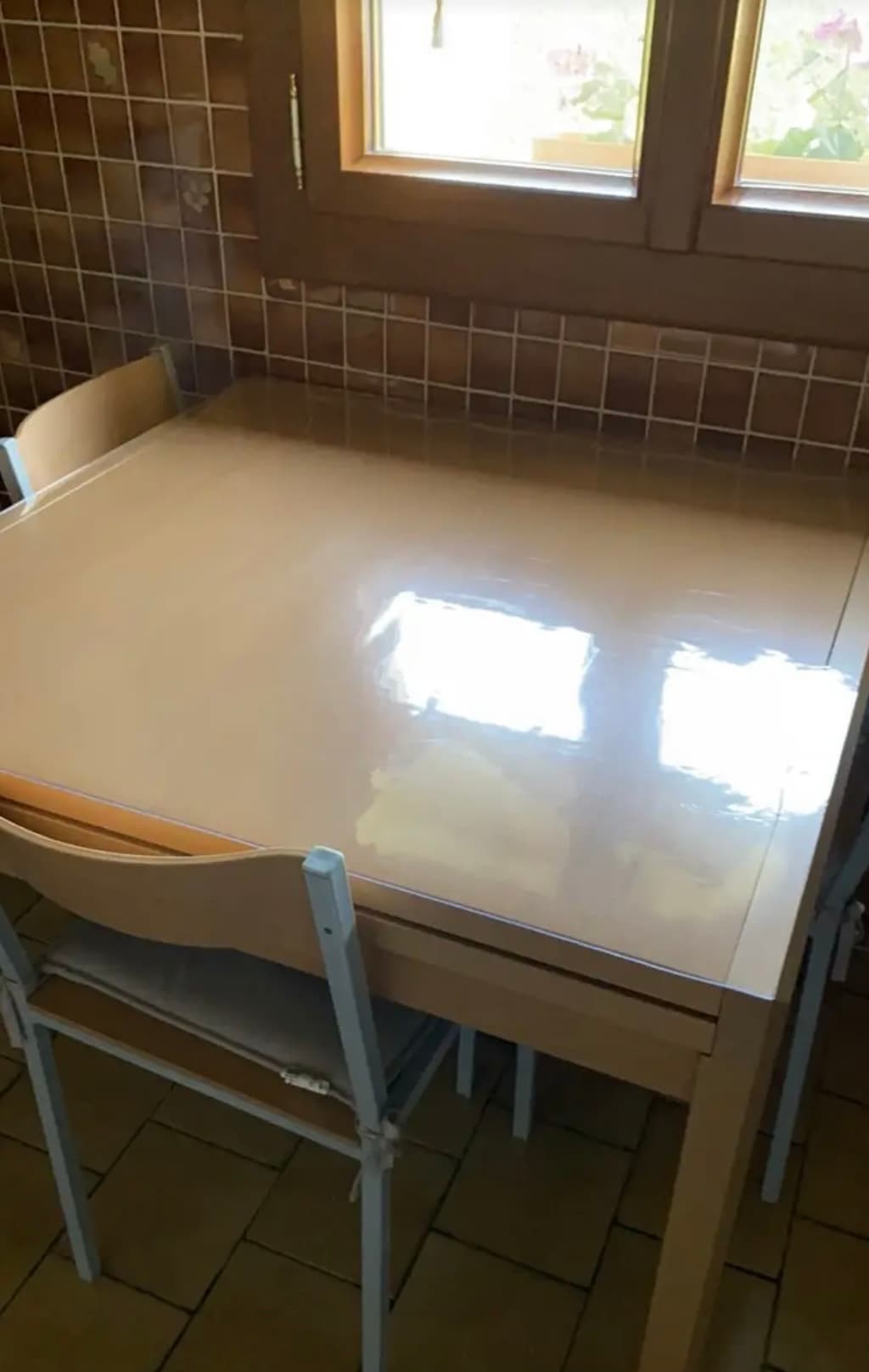 Klein crèmekleurig tafeltje in een betegelde keuken; op het blad de mat, in het raam potten met geraniums