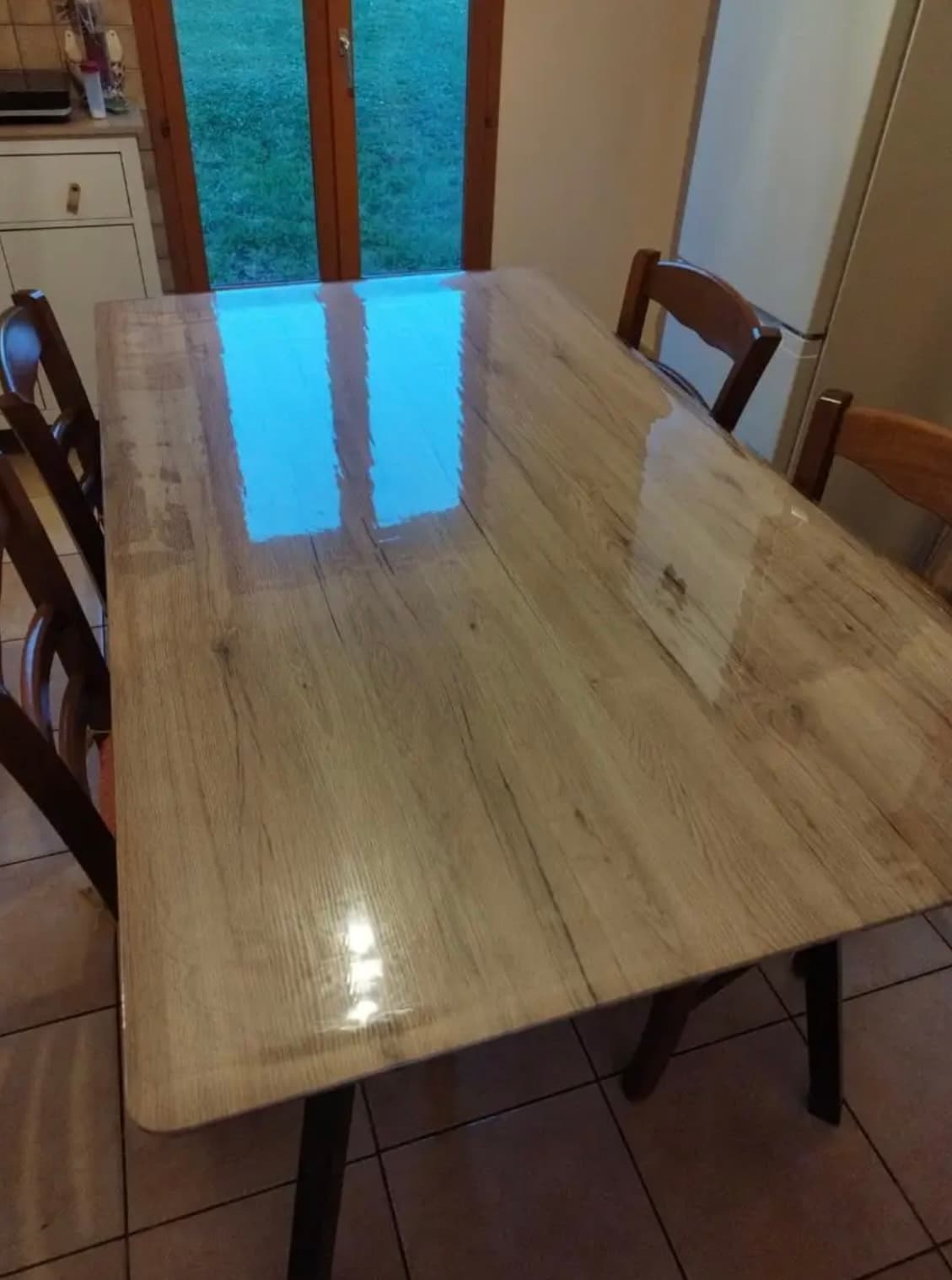 Keukentafel van licht eiken; in de mat weerspiegelt de deur naar de tuin