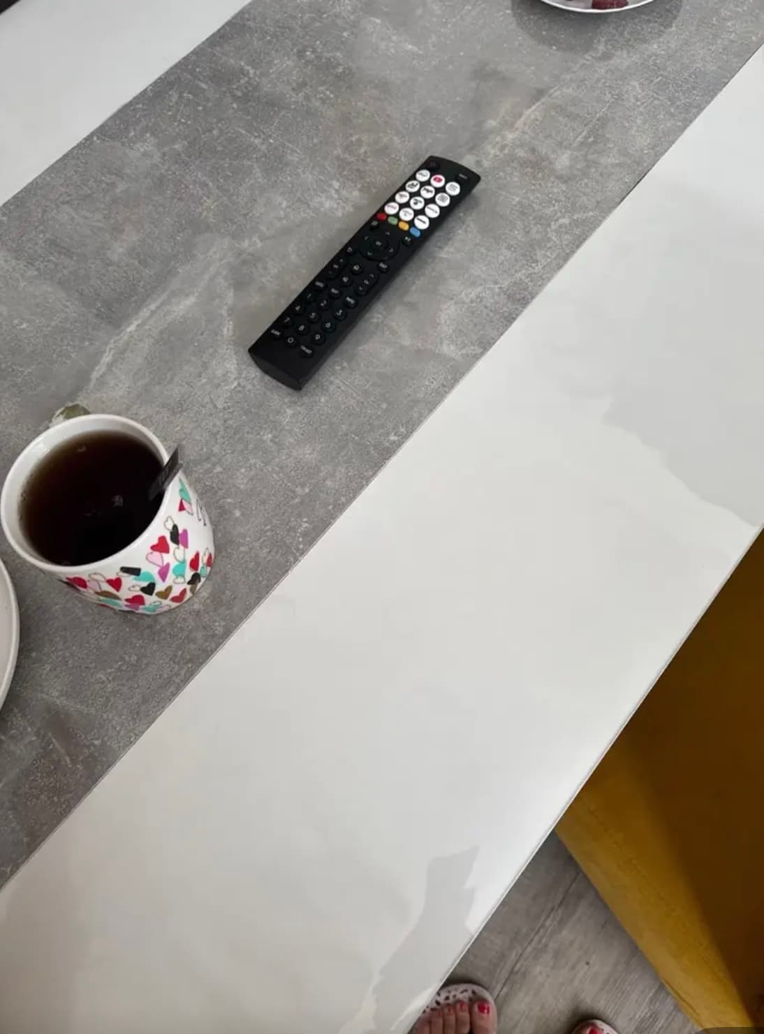 Tafel met wit blad en grijze betonstrook: een kop thee en een afstandsbediening liggen op de mat